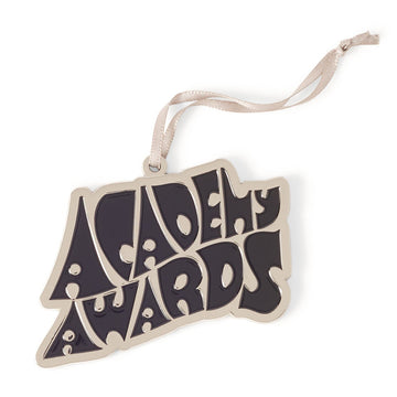 An enamel holiday ornament featuring the text 'Academy Awards' in a stylized font, with a ribbon attachment for hanging.